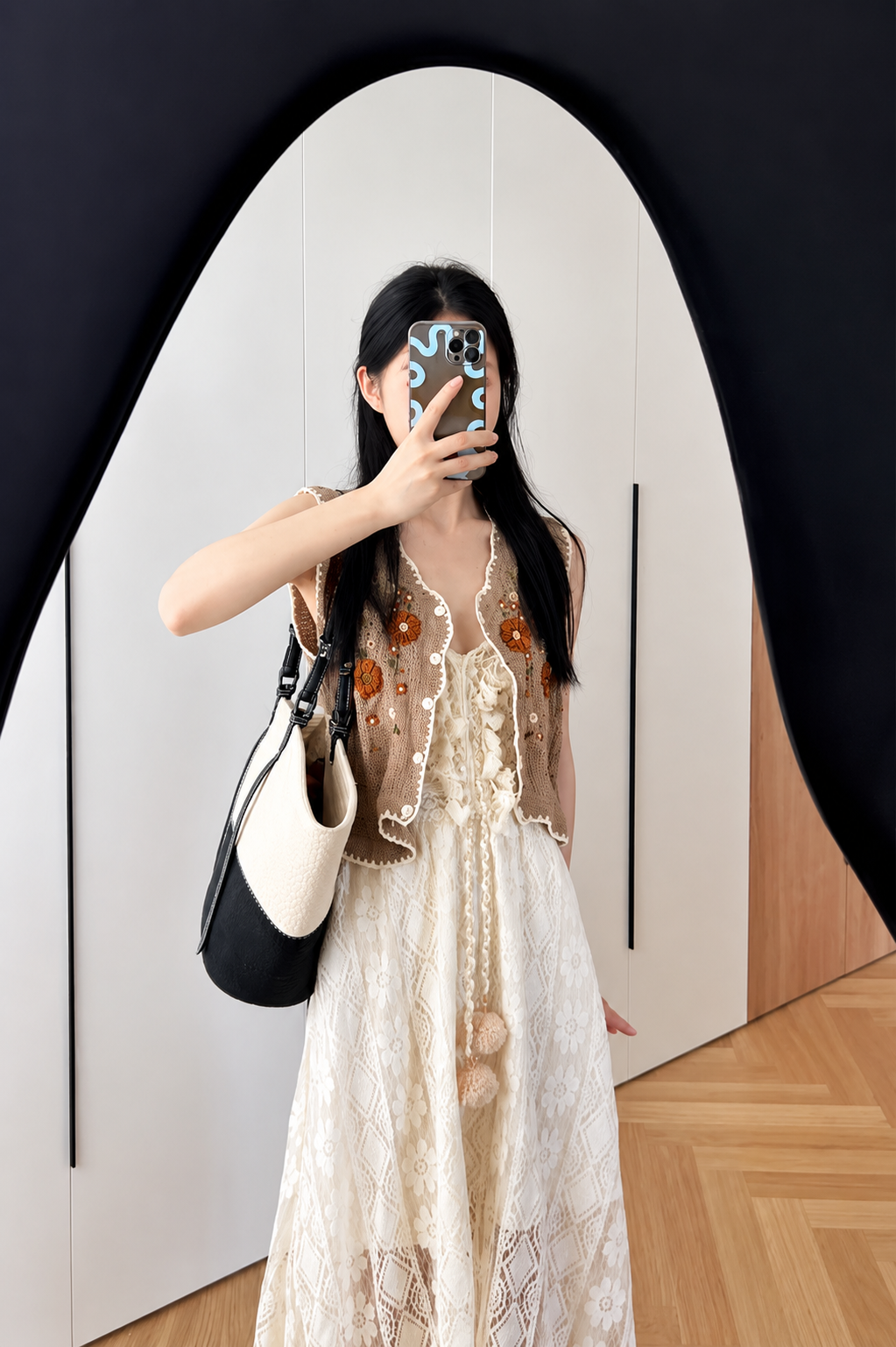 Woman taking a mirror selfie wearing a beige lace top and white skirt with a black and white handbag.