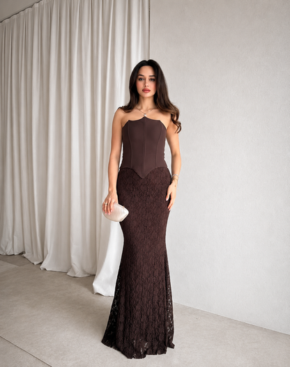 Woman in a dark brown strapless lace evening gown holding a white clutch against a neutral background